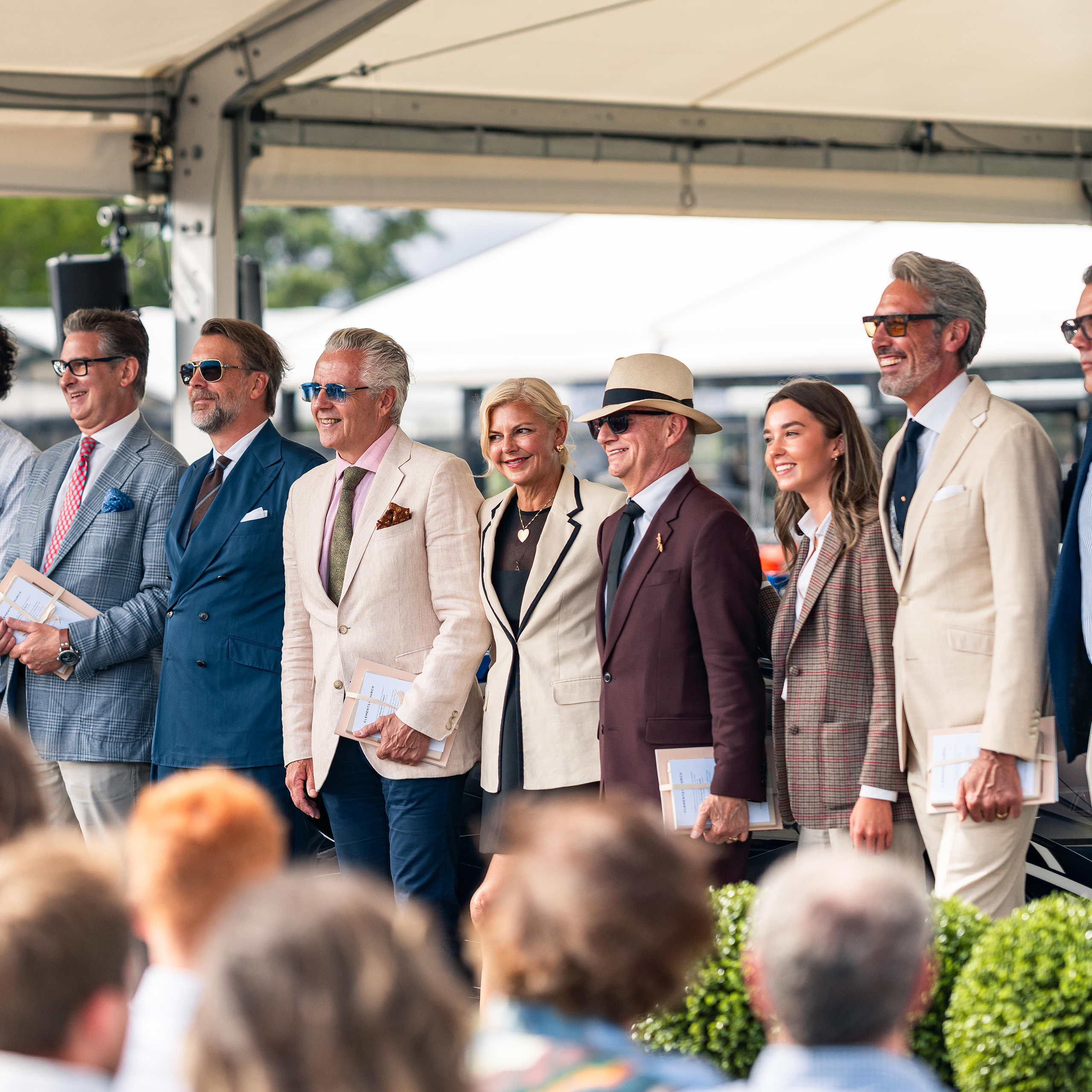 Group of guests in tailored jackets standing on the Concours d'Élégance stage under an event awning