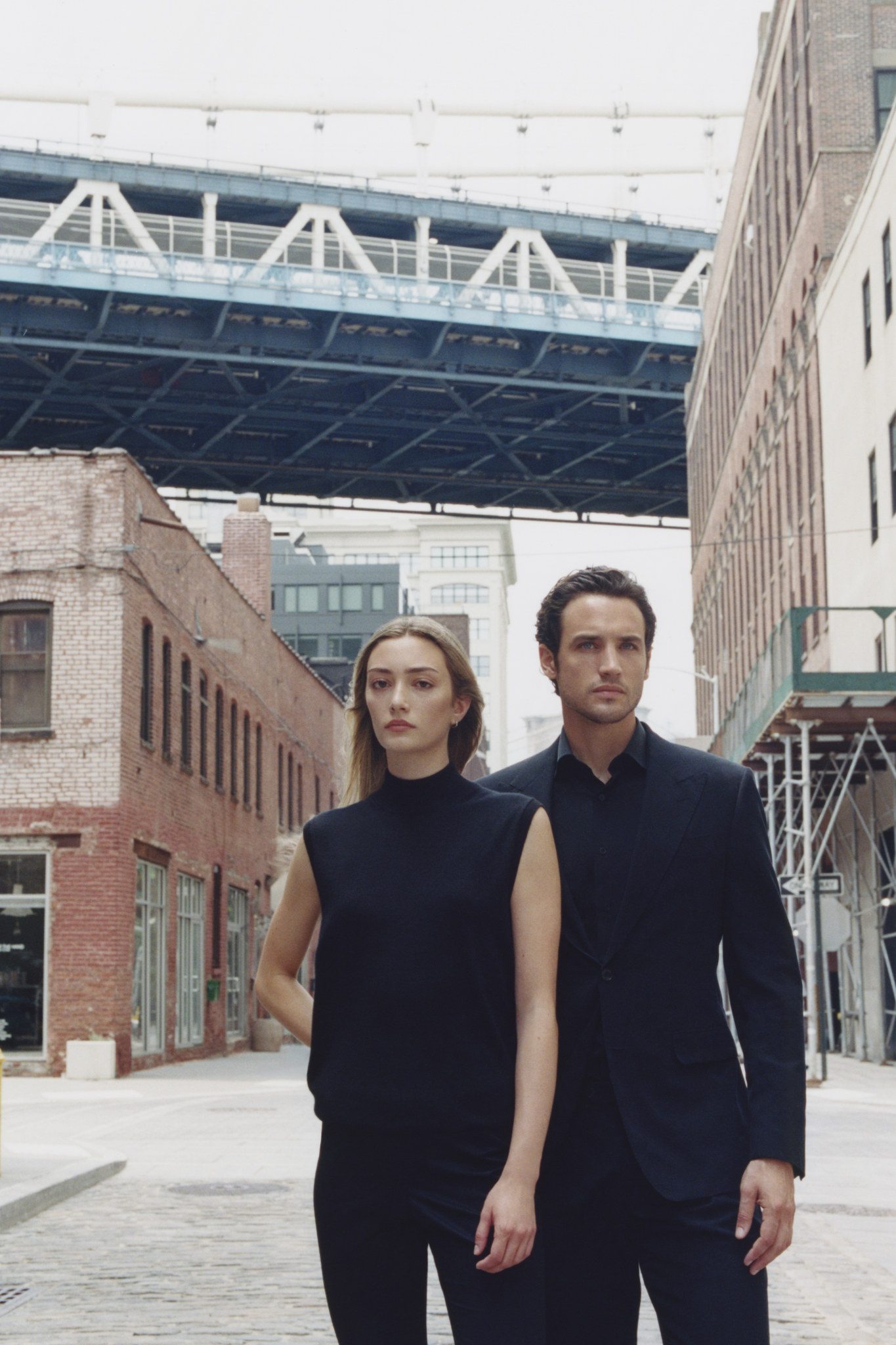 Two models in black Clements+Church tailoring walking beneath the Manhattan Bridge in New York