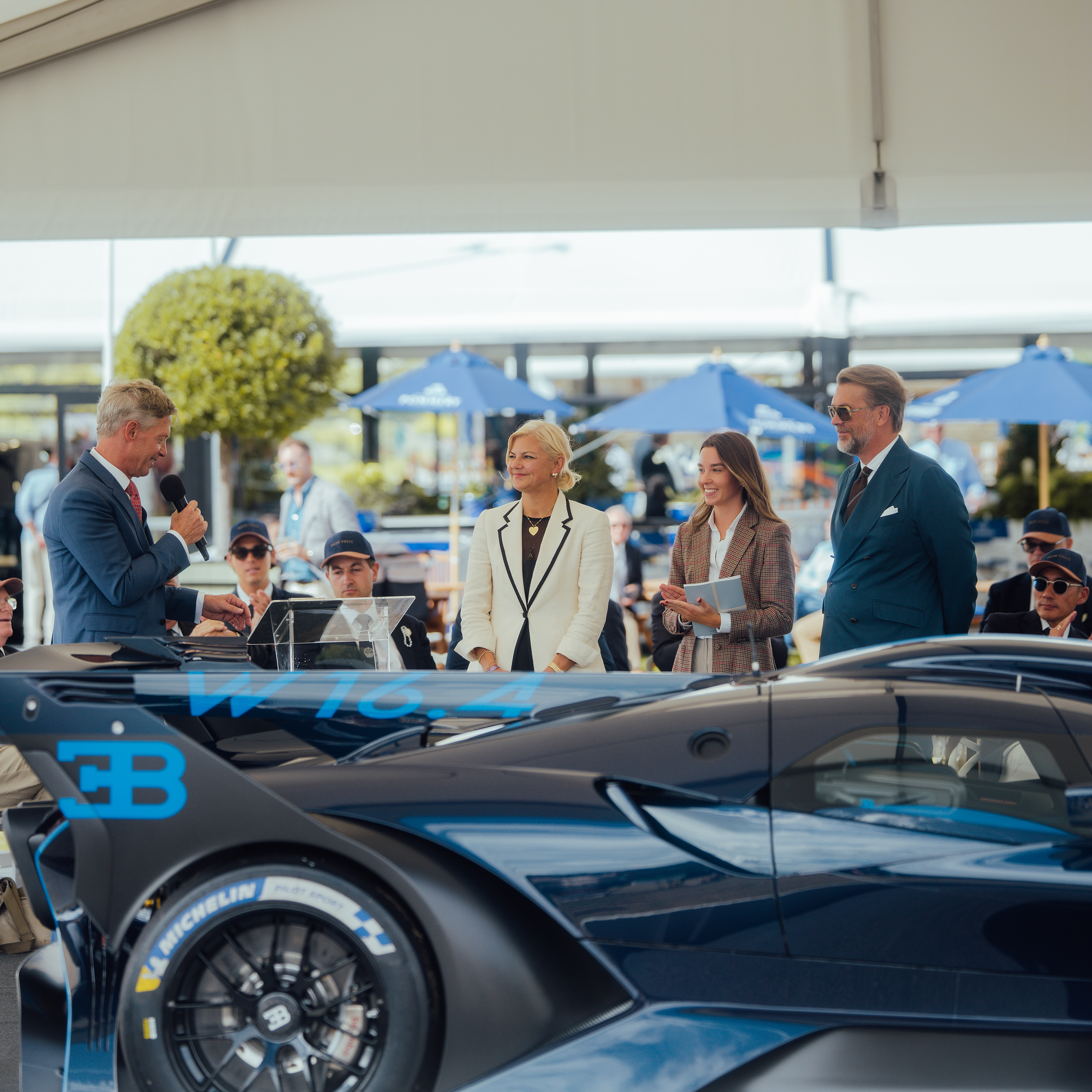 Presenter with microphone alongside guests beside a blue Bugatti at the Concours d'Élégance