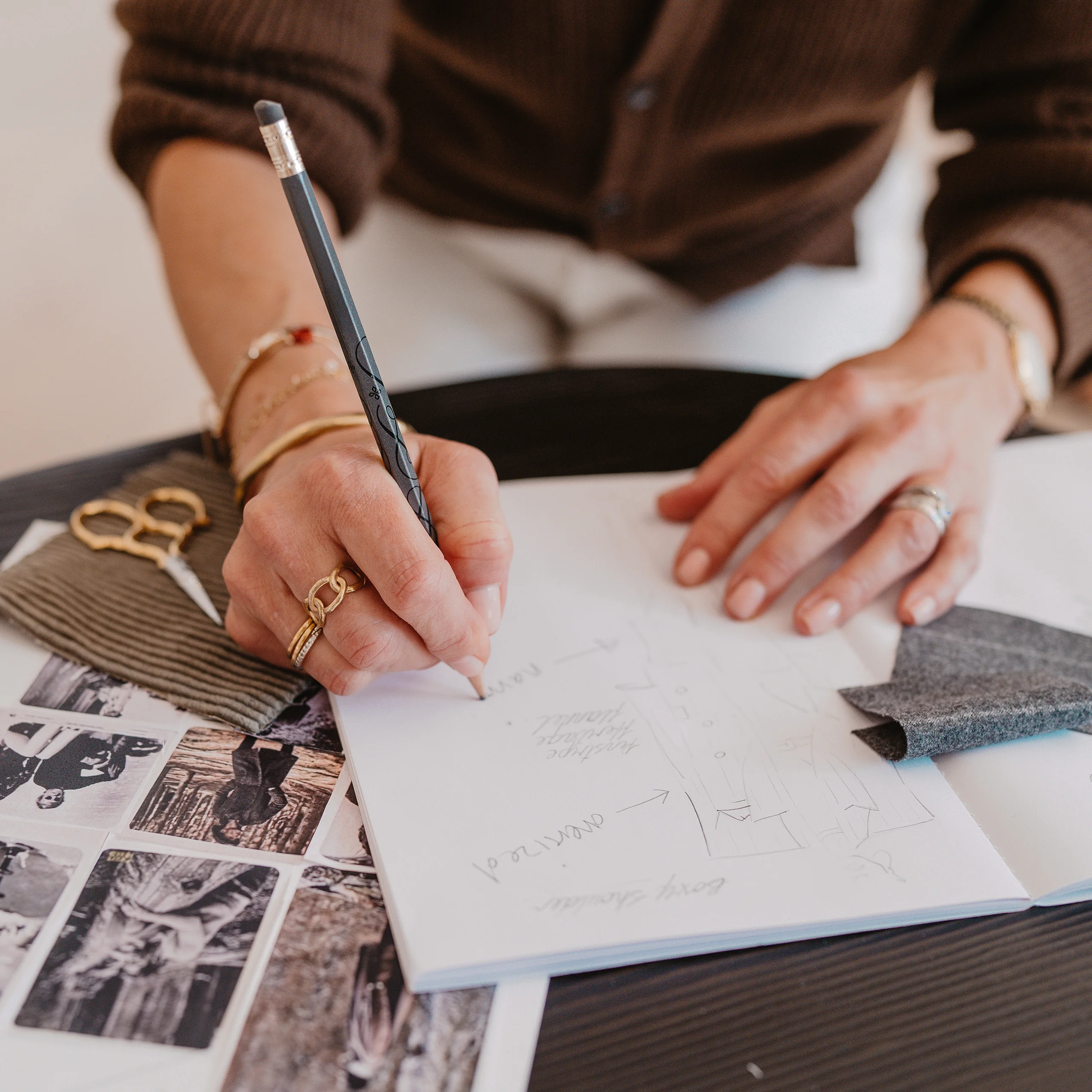 A hand sketching a waistcoat in a notebook, with fabric swatches and gold scissors laid out on a dark timber table.