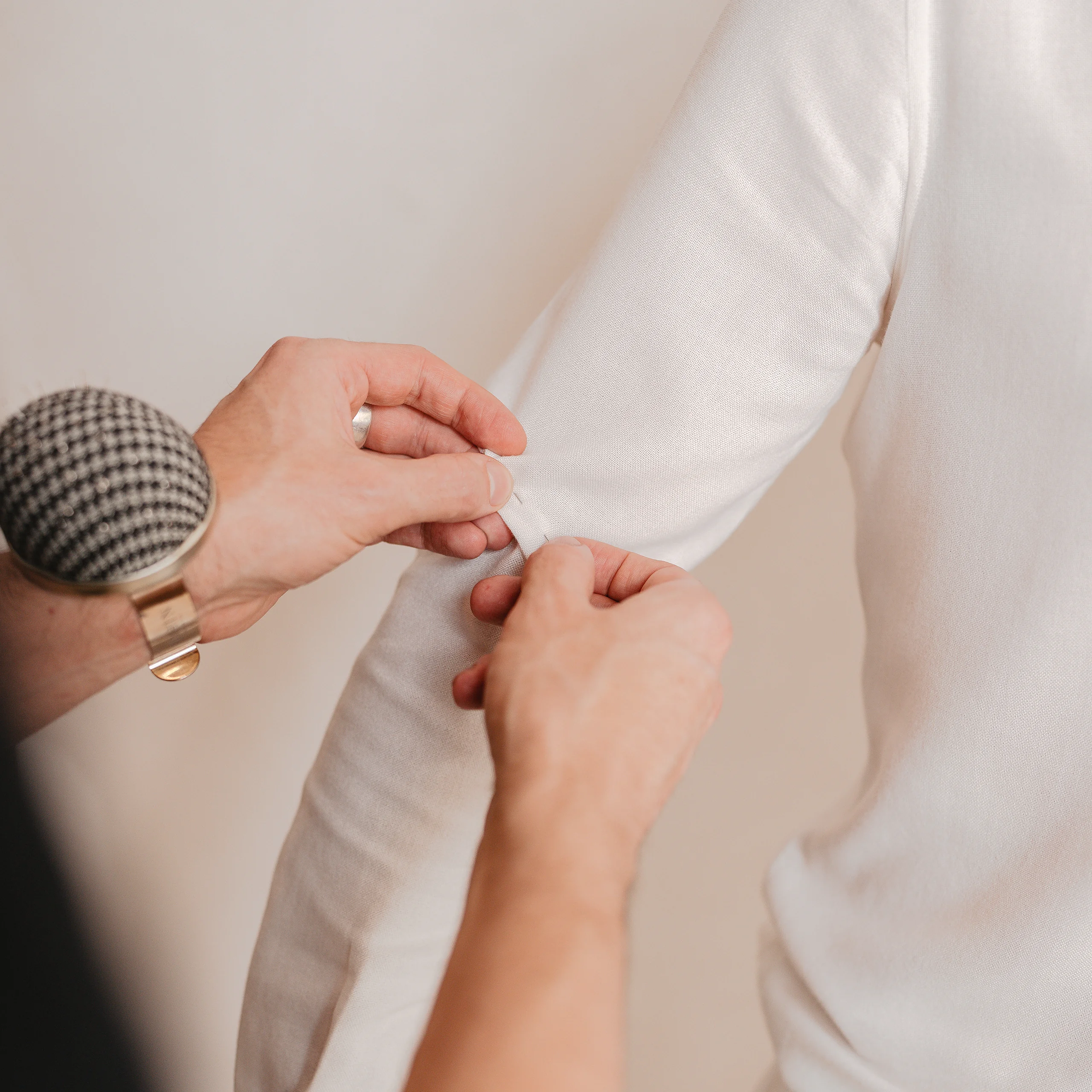 A tailor basting a sleeve seam on a white cotton toile, wearing a patterned wrist pincushion.