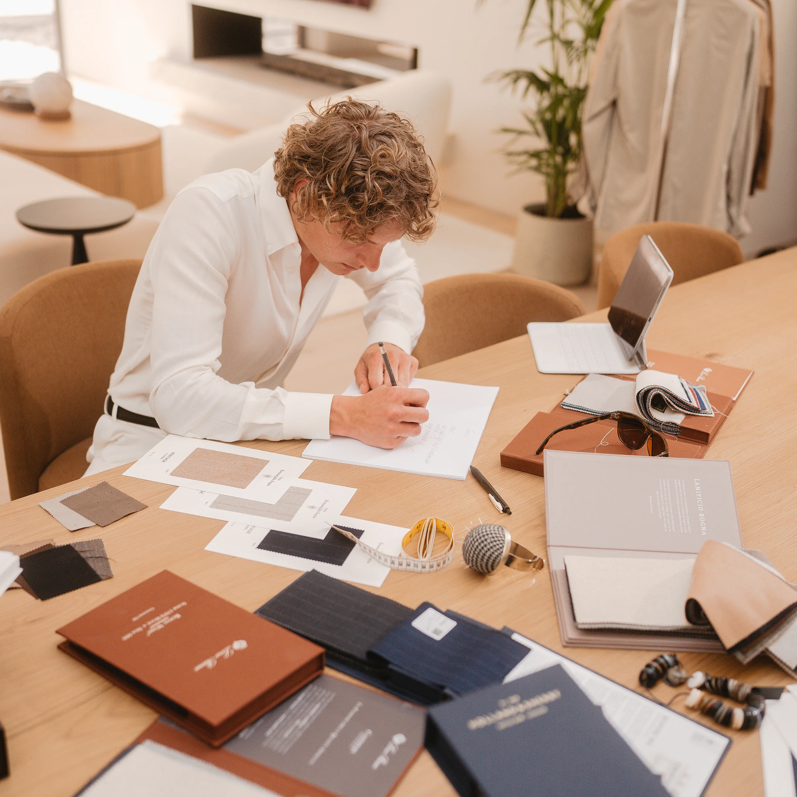 Designer sketching on paper at a light oak desk covered with fabric swatches, notebooks and a tape measure