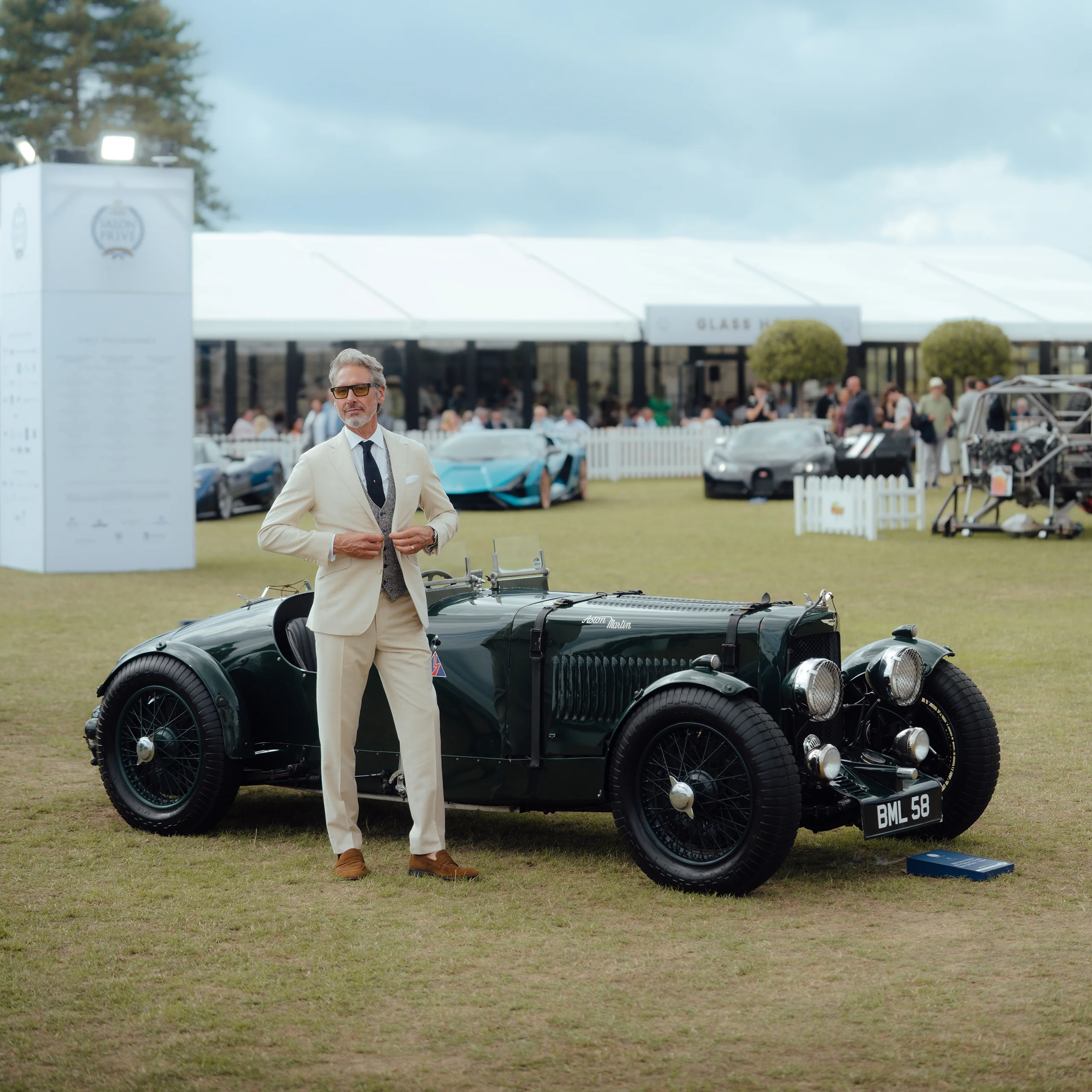 A man in a Clements and Church cream suit standing beside a vintage green Aston Martin at Salon Privé