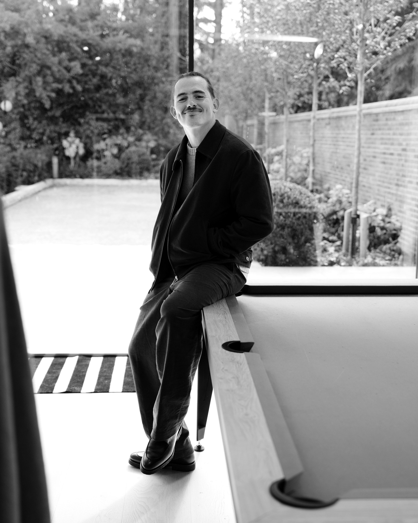 Freddie Grant, Beaconsfield Tailor, seated on the edge of a pool table in a dark jacket and trousers