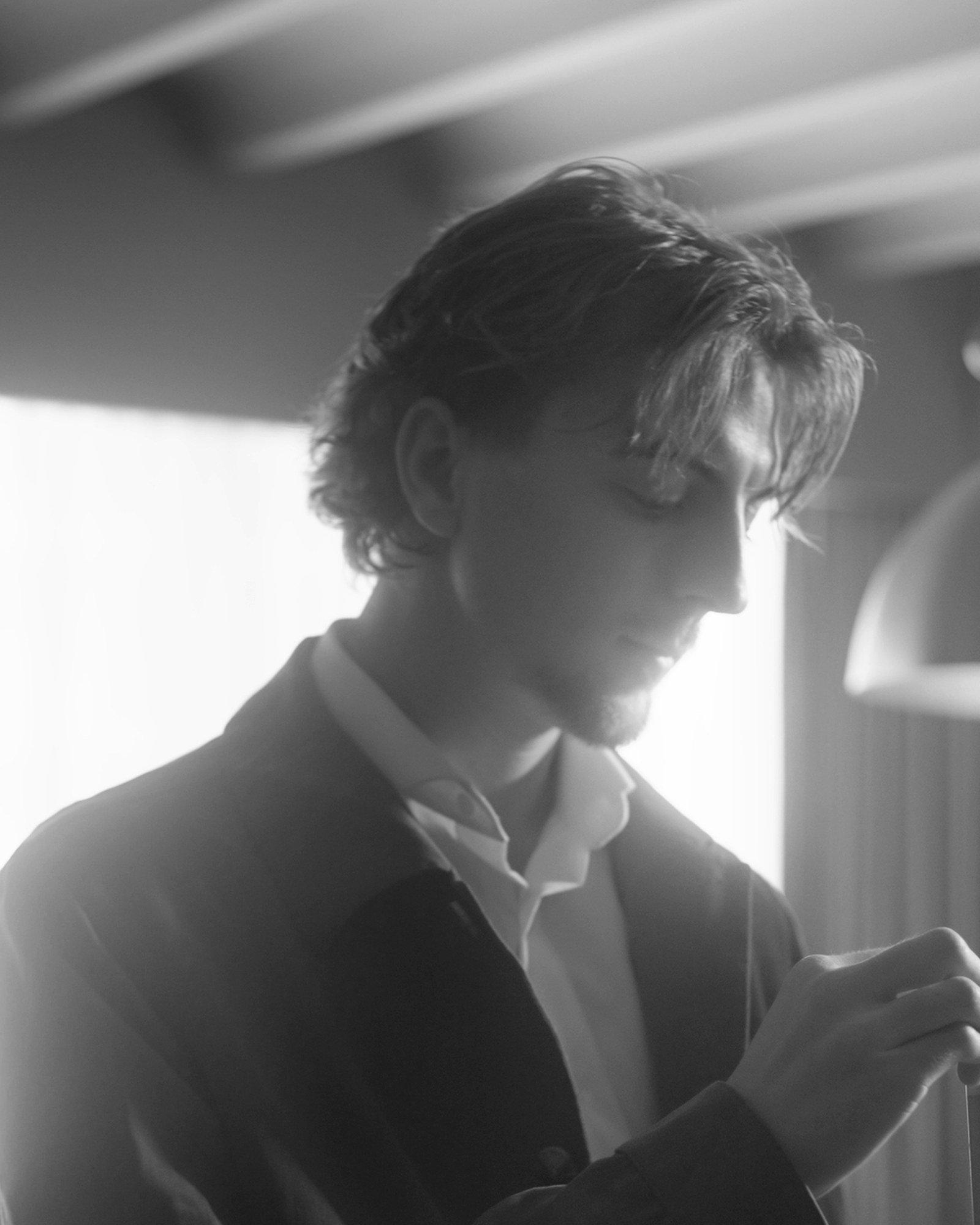 Harry Clements, Dubai Tailor, looking down in soft window light in a dark jacket and open-collar shirt