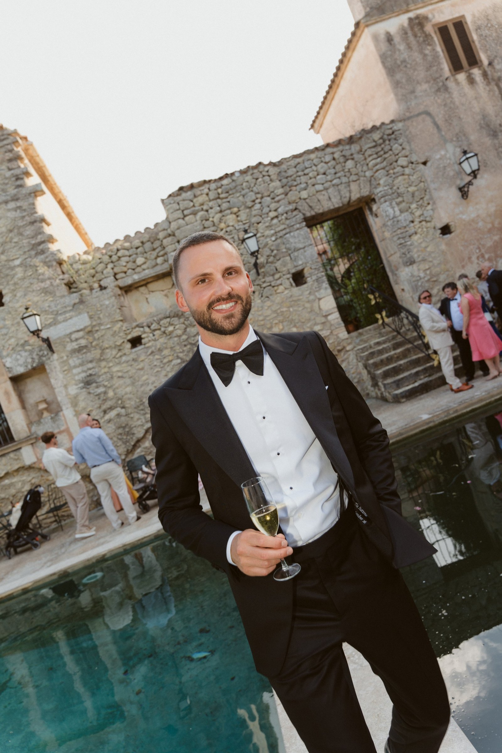 Brad in a black Clements+Church tuxedo and bow tie holding a glass of champagne beside a pool