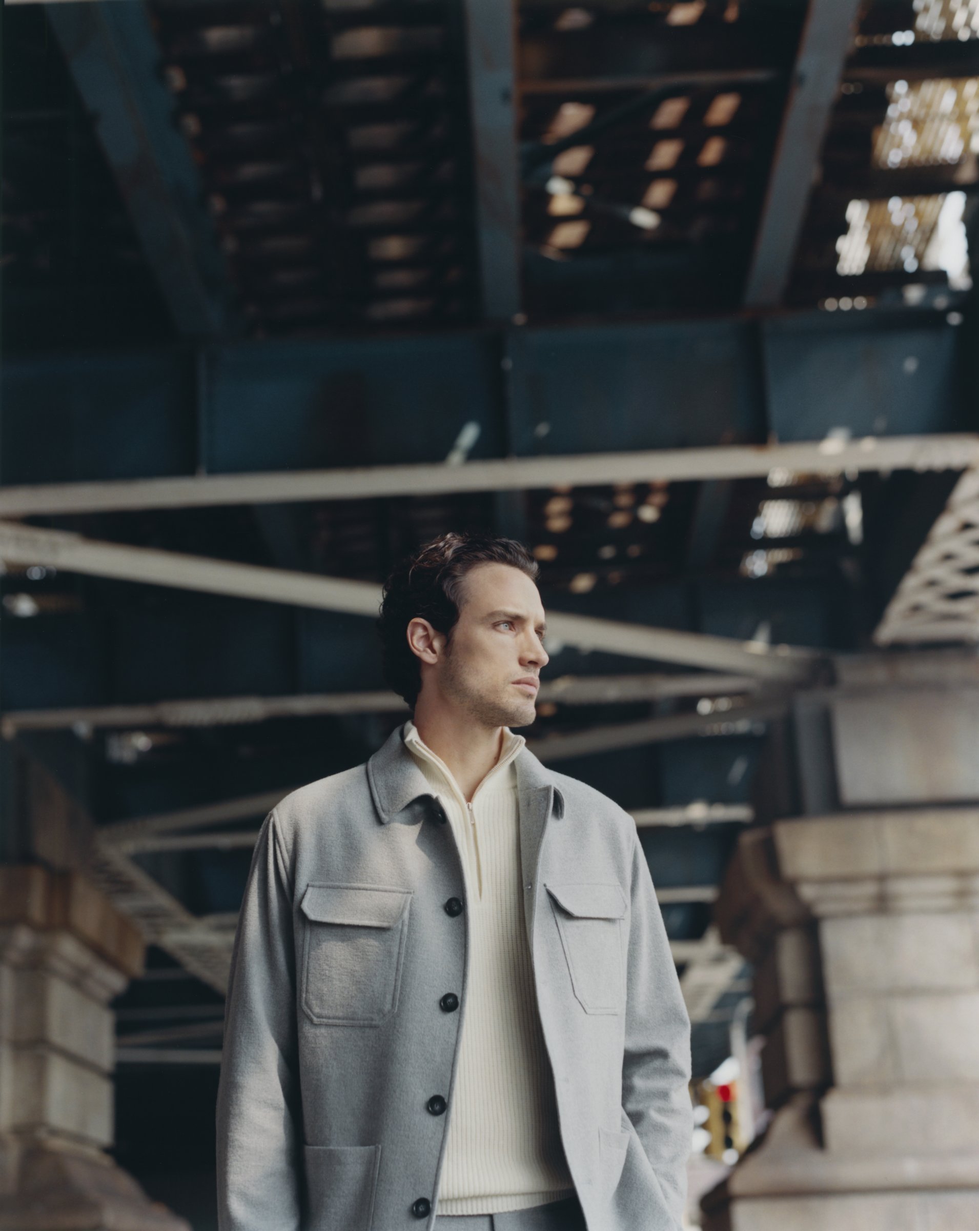 Man in a custom made grey Subalpino winter seersucker chore coat and cream cashmere half-zip beneath a New York bridge